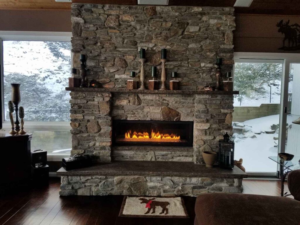 A stone fireplace with a mantel, positioned beneath a large window in a cozy living room setting.